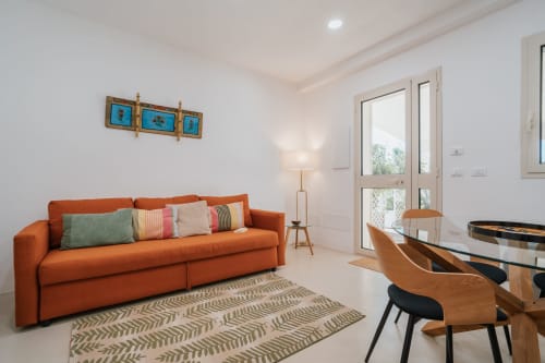 Luminoso soggiorno con divano arancione, tavolo in legno e porte-finestre con vista giardino all'appartamento Case Luparello di Noto