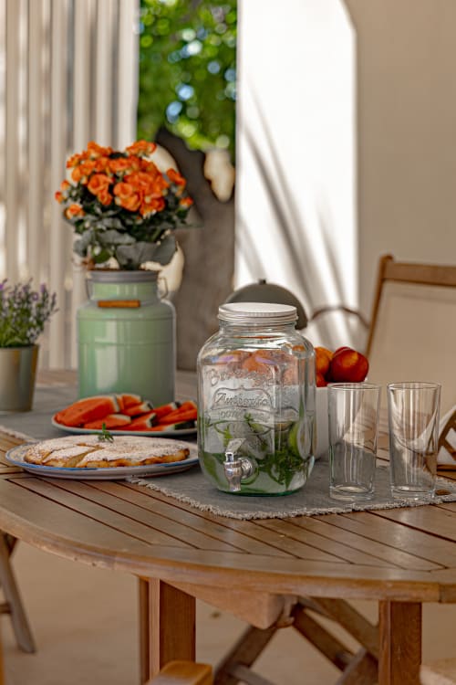 Tavolo da pranzo all'aperto a Case Luparello con verdure fresche, pane, dispenser in vetro vintage e fiori d'arancia con vista panoramica