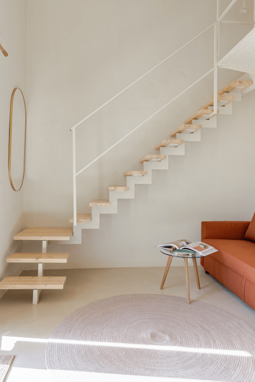 Zona living minimalista moderna a Case Luparello con scale in legno sospese, divano arancio e tappeto circolare a Noto