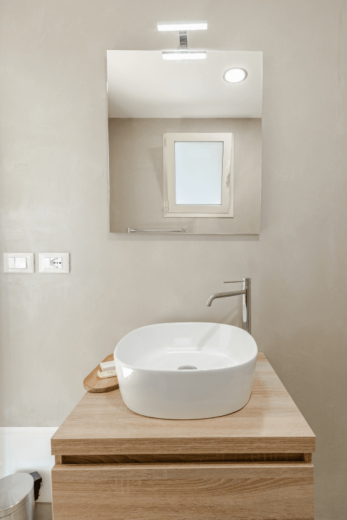 Bagno moderno con lavabo bianco su mobile in legno, specchio e finestra presso l'hotel Case Luparello a Noto