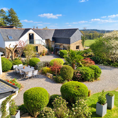 
Vue panoramique du jardin paysager et de la cour fleurie de Ty Coz Breizh, maison d’hôtes à Guidel en Bretagne Sud, avec terrasse, salon de jardin et bâtiments en pierre.
