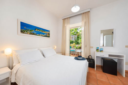 Camera luminosa a Villa Bina Sea Hotel con letto bianco, balcone con vista giardino e opere d'arte costiera a Serrara Fontana