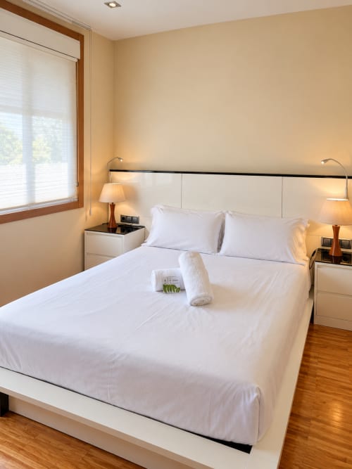 Dormitorio moderno en apartamento Aires de Baiona, Baiona con ropa de cama blanca, mesitas de noche gemelas con lámparas y suelo de madera
