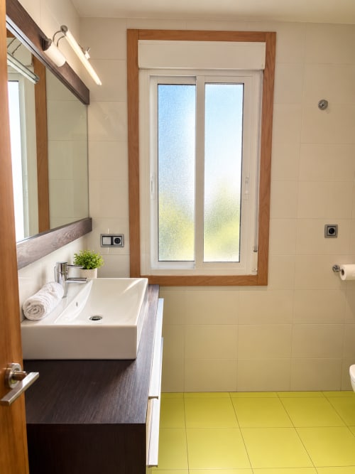 Baño moderno en apartamento de Baiona con lavabo tipo vasija, espejo con marco de madera, suelo de azulejos amarillos y puertas de cristal esmerilado