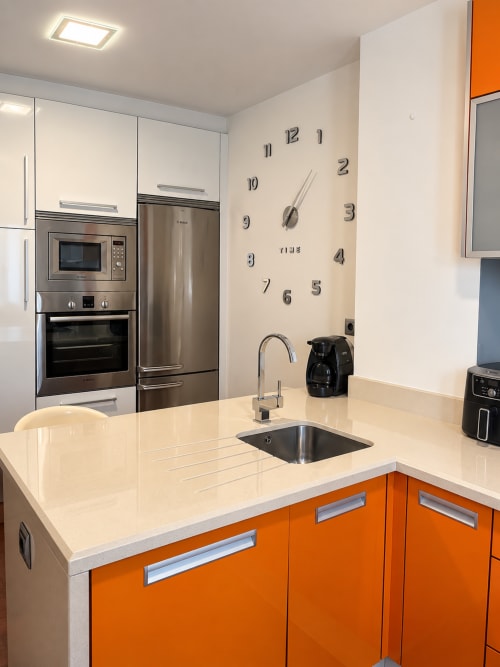 Cocina moderna en apartamento de Baiona con muebles naranjas, encimera blanca, electrodomésticos de acero inoxidable y reloj de pared