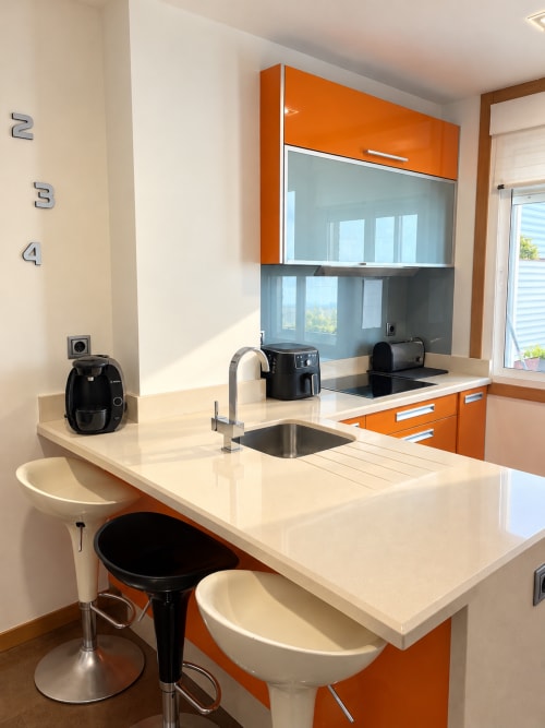 Cocina moderna en apartamento de Baiona con gabinetes naranjas, encimeras blancas, fregadero de acero inoxidable y asientos de bar
