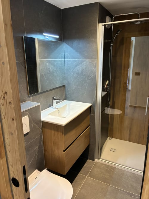 Salle de bain moderne avec meuble vasque en bois, lavabo blanc, murs carrelés sombres et douche vitrée aux Tanières de Montgenèvre