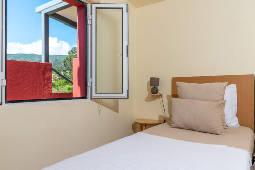 Bright bedroom at Garry's Holiday Rentals in Prazeres with wooden bed, beige pillows, and windows overlooking green hills and