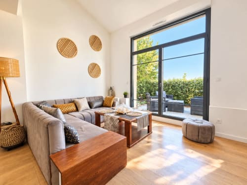 Sala de estar moderna con sofá gris, mesas de madera y grandes ventanales con vistas a los jardines de La Perle du Léman