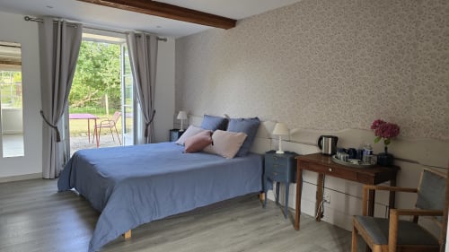 Bright bedroom with blue bedding, garden views through glass doors, wooden beams, and modern amenities at Domaine de la Cour