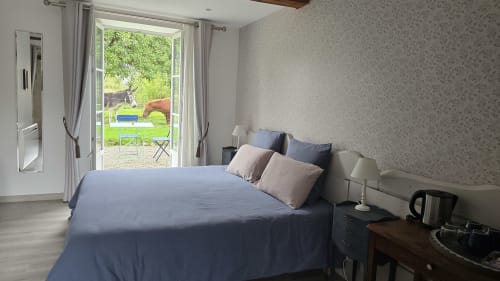 Chambre lumineuse avec literie bleue, portes-fenêtres sur terrasse avec vue jardin et papier peint floral au Domaine de la Cour VAUTIER près d'Omaha