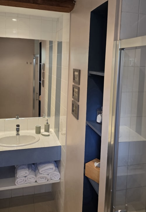 Modern bathroom with white tile walls, navy blue vanity with sink, rolled towels, and glass shower enclosure at Domaine de la