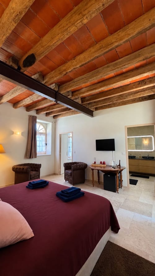 Spacious bedroom at Cloffy B&B with exposed wooden beams, burgundy bed, leather seating, and TV in Nouans-les-Fontaines