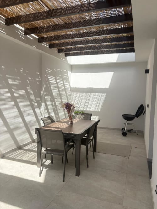 Sala de jantar moderna com pérgola de listras, luz natural, mesa com cadeiras e flores em vasos na villa BHF Residences