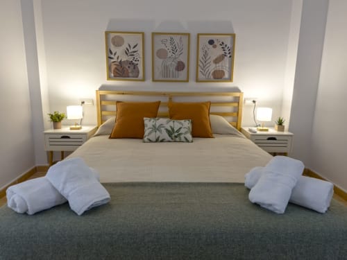 Dormitorio moderno en Living Sur cottage, Sanlúcar de Barrameda, con cama de madera, detalles dorados, toallas blancas y pared botánica