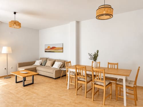 Moderna sala de estar y comedor del apartamento Alzara en Sanlúcar de Barrameda con sofá beige, muebles de madera y vistas a la montaña