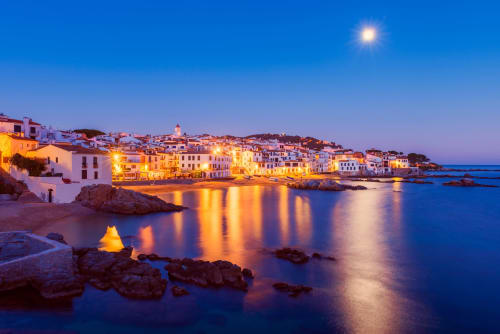 Pueblo costero de Llafranc al atardecer con edificios iluminados reflejándose en aguas azules tranquilas y orilla rocosa