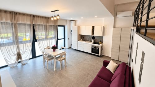 Zona living di un moderno appartamento all'hotel Saronno con divano bordeaux, angolo cottura, tavolo da pranzo e vista sulla città