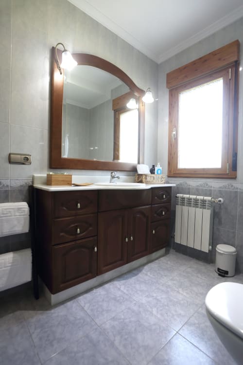 Baño espacioso de A Quinta de Lapido con tocador de madera oscura, espejo, lavamanos blanco y piso de baldosas