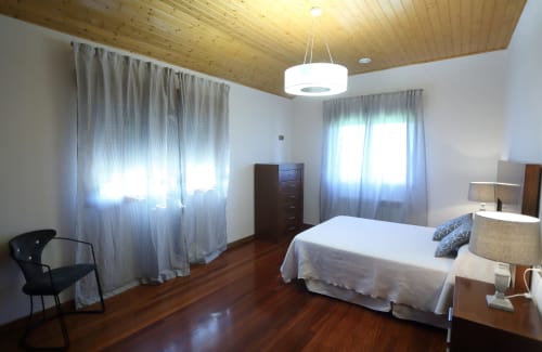 Dormitorio luminoso y espacioso en A Quinta de Lapido con techo de madera, dos ventanas, cama queen, cómoda y lámpara colgante moderna