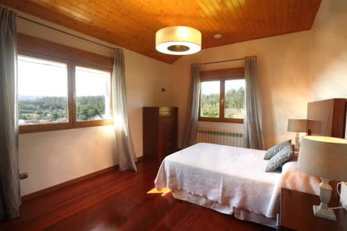 Dormitorio luminoso en A Quinta de Lapido con techo de madera, dos grandes ventanales con vistas a árboles, cama blanca y calidez en tonos cálidos