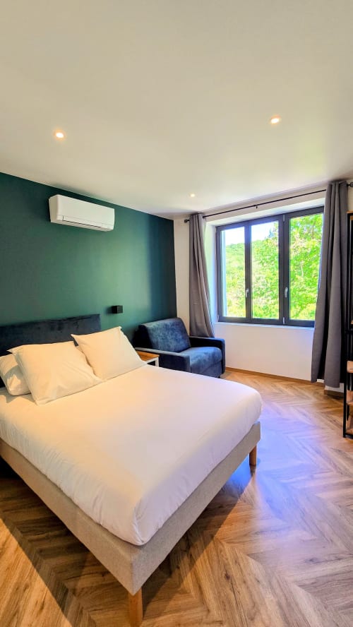Modern bedroom with green accent wall, white bed, armchair, large windows overlooking greenery at Vindihouses Épinac BnB