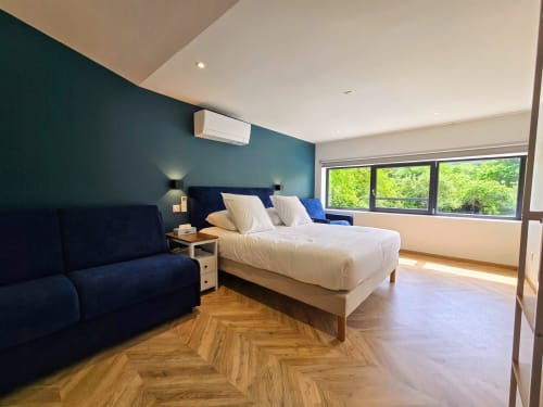 Modern bedroom at Vindihouses in Épinac with teal accent wall, white bedding, dark blue sofa, and large windows overlooking