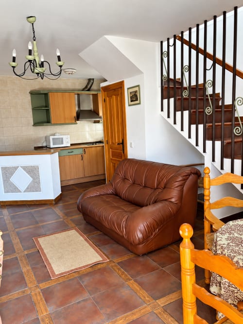Apartamento de planta abierta con sofá de cuero marrón, escaleras de madera con barandillas de hierro forjado, cocina integrada y suelos de baldosa