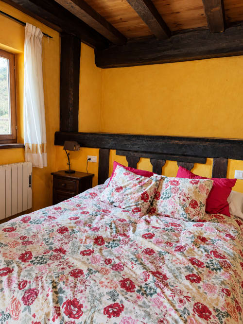 Dormitorio rústico con paredes amarillas, vigas de madera oscura y ropa de cama floral en Casa rural El Toleo en Hoyos del Espino