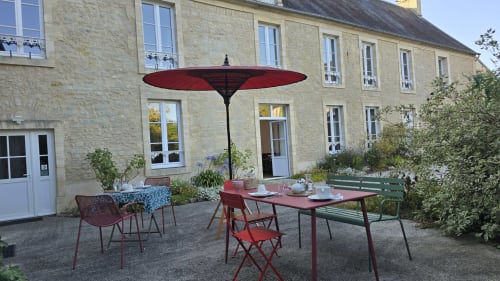 Terrasse du jardin de Domaine de la Cour VAUTIER avec parasol rouge, table à manger et maison de pierre à Mandeville-en-Bessin
