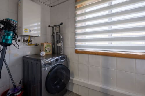 Laundry room with black washing machine, white tiled walls, window with blinds, and hanging bicycle at EcoHost apartment
