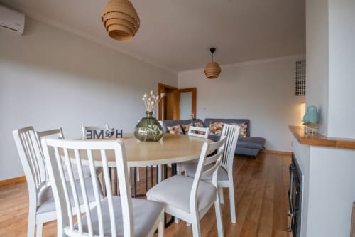 Modern dining area with white chairs at round table, living room with gray sofa, wooden floors, and pendant lights in