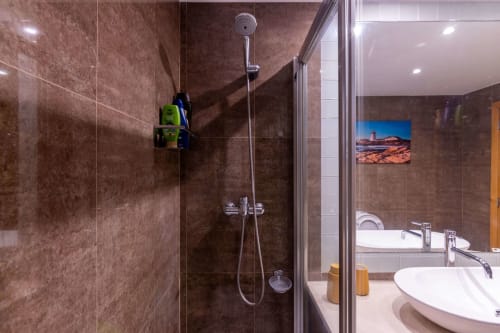 Modern bathroom shower in EcoHost Matosinhos apartment with brown tiled walls, rainfall showerhead, and coastal view artwork.