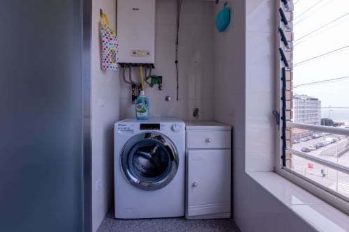 Compact laundry corner in EcoHost apartment with washing machine, dryer, wall-mounted boiler, and sea view window overlooking
