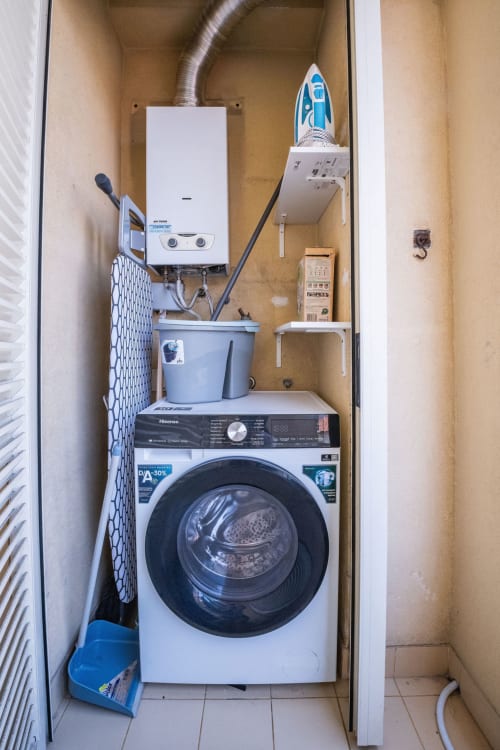 Compact laundry closet in EcoHost apartment with Hisense washing machine, white gas water heater, ironing board, and storage