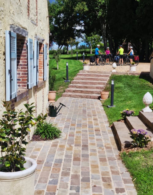 Cour de la Casa Pertica à Ceton avec cyclistes, jardins soignés, allées en briques et architecture historique en pierre