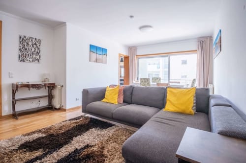 Modern living room with gray sectional sofa, yellow pillows, wood flooring, area rug, and large windows at EcoHost apartment