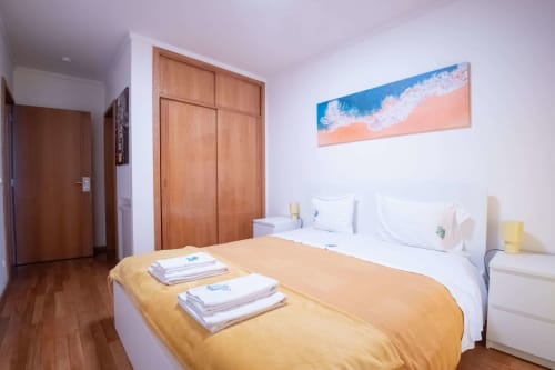 Bright bedroom at EcoHost Gaia Beach&Golf in Arcozelo with queen bed, yellow throw, beach artwork, wooden wardrobe, and wood