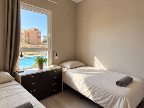 Bright shared hostel bedroom at Banama Coliving in Corralejo with twin beds, pool view window, black desk, and modern