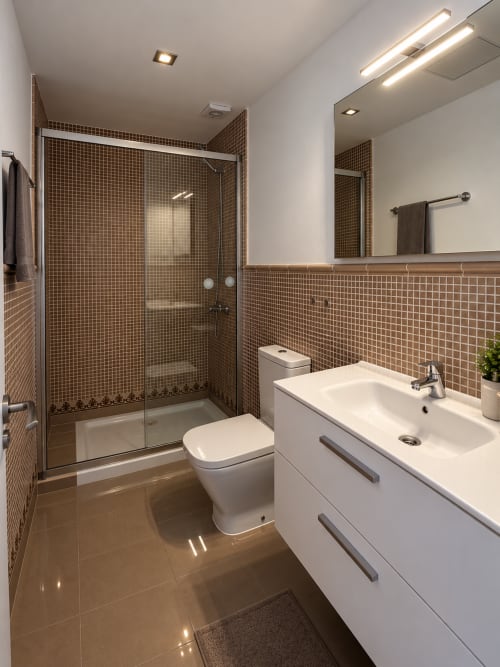 Modern bathroom with brown mosaic tiles, white vanity, toilet, and glass shower enclosure at Banama Coliving hostel in