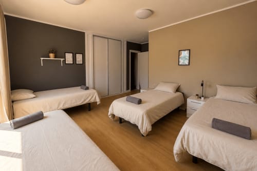 Shared 4-bed room at Banama Coliving hostel in Corralejo with cream bedding, wooden floors, and dark accent wall.