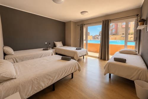 Bright 4-person shared room at Banama Coliving Corralejo with beige beds, dark accent wall, pool view balcony, and wooden