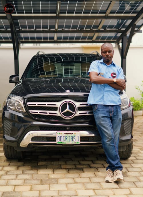 Property manager at OD Cribs Abuja standing with arms crossed beside black Mercedes SUV with solar carport backdrop