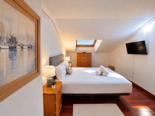 Dormitorio luminoso en ático de Baiona con tragaluz, cuadro de vista marina, cómoda de madera y televisor de pantalla plana