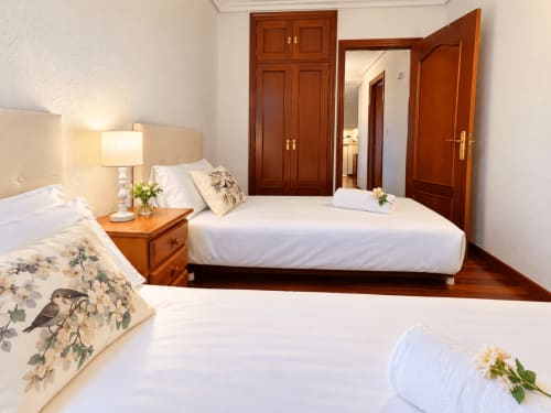 Luminoso dormitorio en apartamento de Baiona con dos camas, mesilla de noche de madera, lámpara y puertas tradicionales de madera