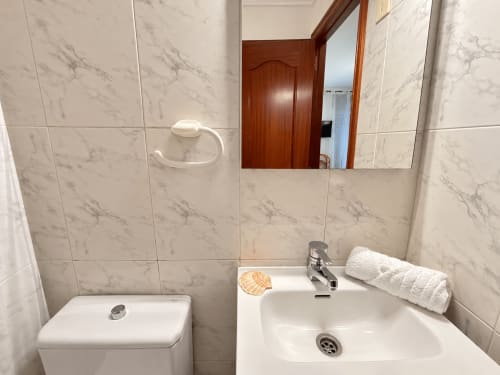 Baño moderno con baldosas de mármol, lavamanos blanco, espejo y toallas en el apartamento Holibai de Baiona, España