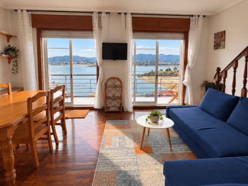 Luminoso salón en Baiona con vistas al mar, sofá azul, mesa de comedor de madera y balcón con vista al agua