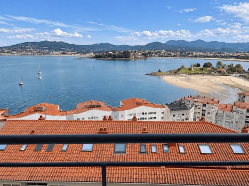Vista de la terraza del apartamento de Baiona con bahía azul, veleros, playa arenosa y montañas