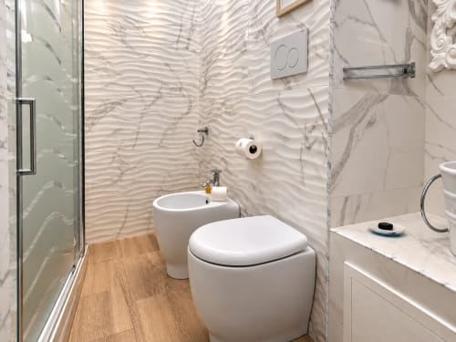 Banheiro moderno da B&B I Mori Catania com vaso sanitário branco, bidê, paredes de mármore, piso de madeira e box de vidro