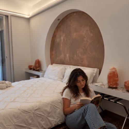 Mujer leyendo en la cama en una acogedora B&B moderna de Catania con cabecero ovalado marrón y lámparas de sal del Himalaya